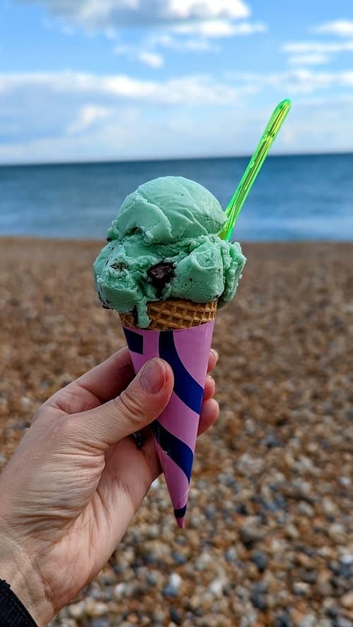ice cream cone filled with one scoop of mint green mint chocolate chip with a neon green spoon held in front of the brighton seafront