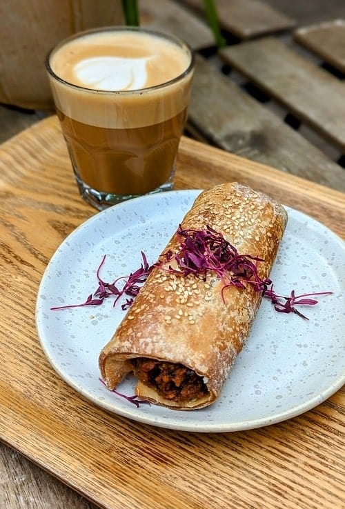 a golden brown vegan and gluten sausage roll on a blue plate next to a small coffee on a wood table at wave in london