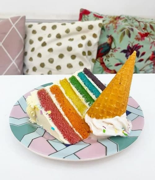 a vegan rainbow cake slice on a colorful plate next to a vanilla ice cream cone that has been flipped on its head at vida bakery in london