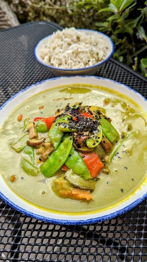 a large bowl of vegan green curry topped with fresh peas, mushrooms, and peppers with a bowl of white rice in the background in london
