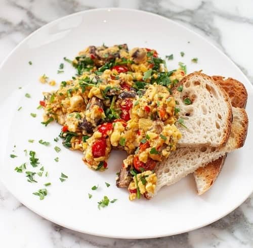 a white plate on a marble counter with a pile of tofu scramble with green and red veggies and toast in london