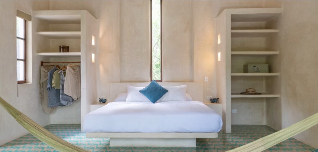 Bright guest room at Holistika Tulum with a king bed, open shelving, colorful tile floors, and a hammock