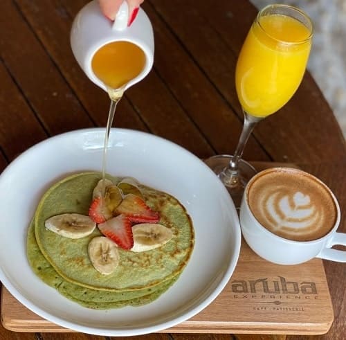 a white plate with two green vegan pancakes topped with banana and strawberries with syrup being poured over them in aruba