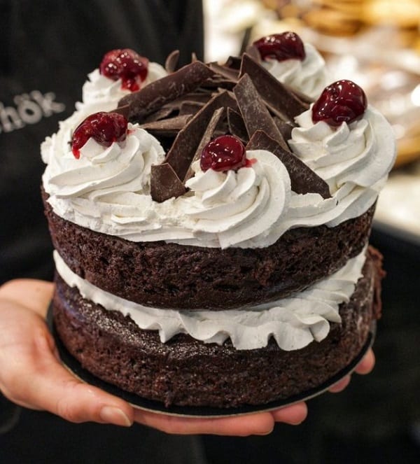 a small vegan chocolate layer cake with white cream in the middle and on top and topped with cherries and chocolate