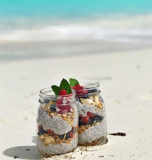 two glass mugs with layers of granola, fruit, and chia seed pudding on the beach in aruba