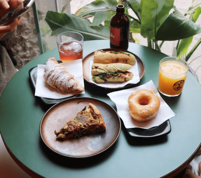 a light blue table filled with multiple dishes that include a vegan slice of cake, croissant, bagel sandwich and juice in barcelona