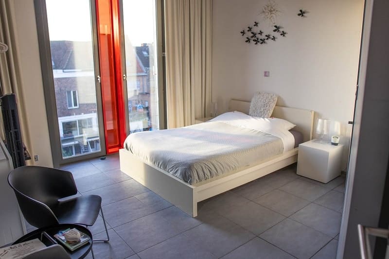 inside of a simple and modern guestroom at vegan b and b in bruges with grey linen bed, white walls, tile floor, and large sunny window