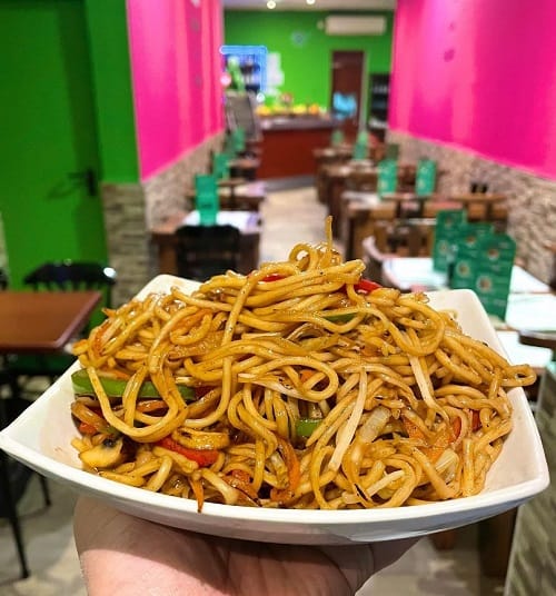 a large bowl of vegan asian noodles held in front of a bright pink and green background at vegan mount in barcelona