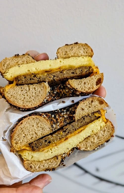 a vegan everything bagel sandwich with sausage and egg cut in half and held in front of a white wall at ben and esther's