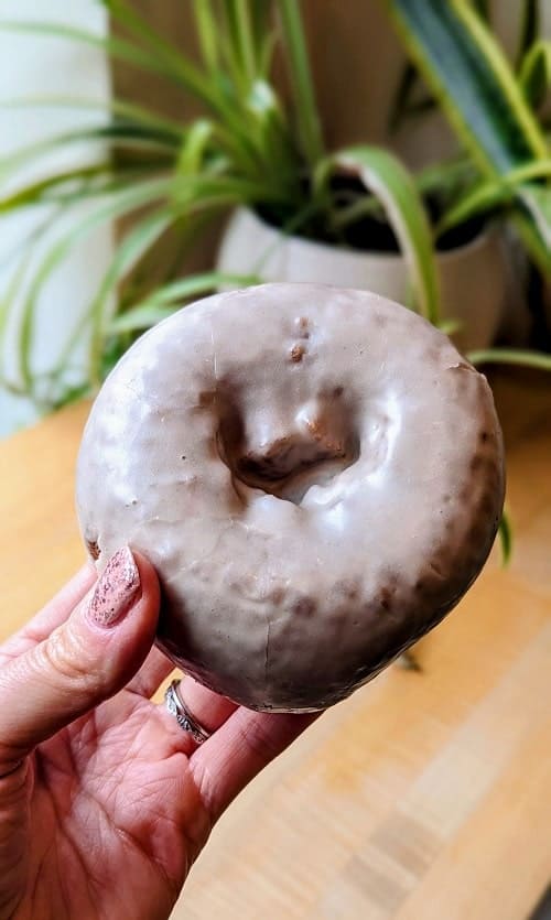 one vegan cake donut covered in a white chai glaze held above a wood table with a plant in the background in portland