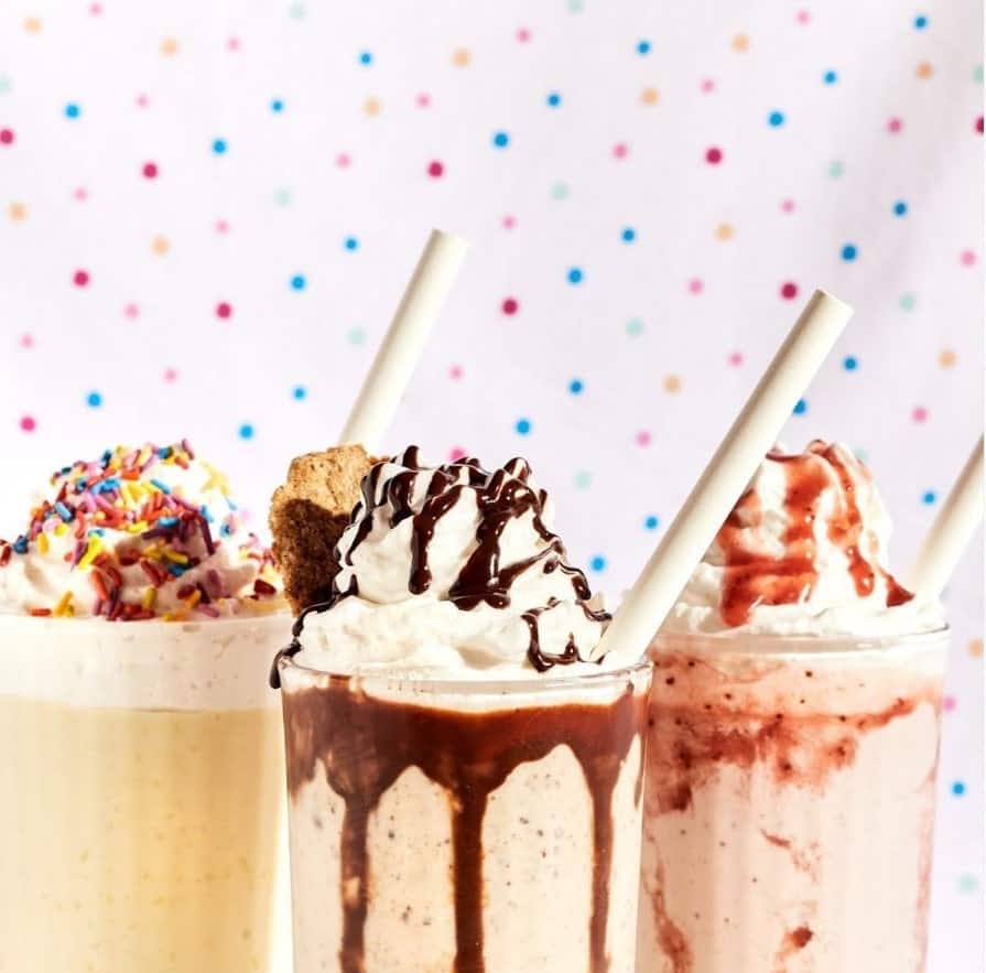 three vegan milkshakes in glass cups next to eachother and topped with whipped cream, chocolate sauce, strawberry sauce, and nuts with a colorful polka dot background behind them