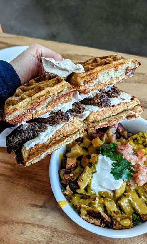 a vegan sausage and cheese waffle sandwich held above a vegan burrito breakfast bowl in portland