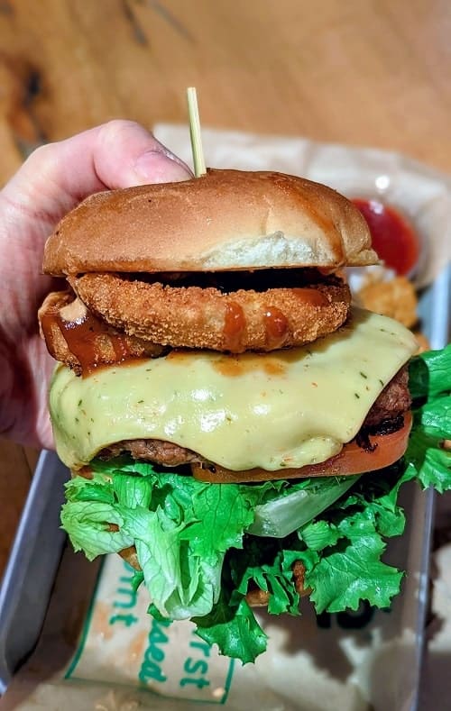 giant vegan burger loaded with melted cheese, lettuce, and a golden onion ring held with one hand over a tray at next level burger in seattle