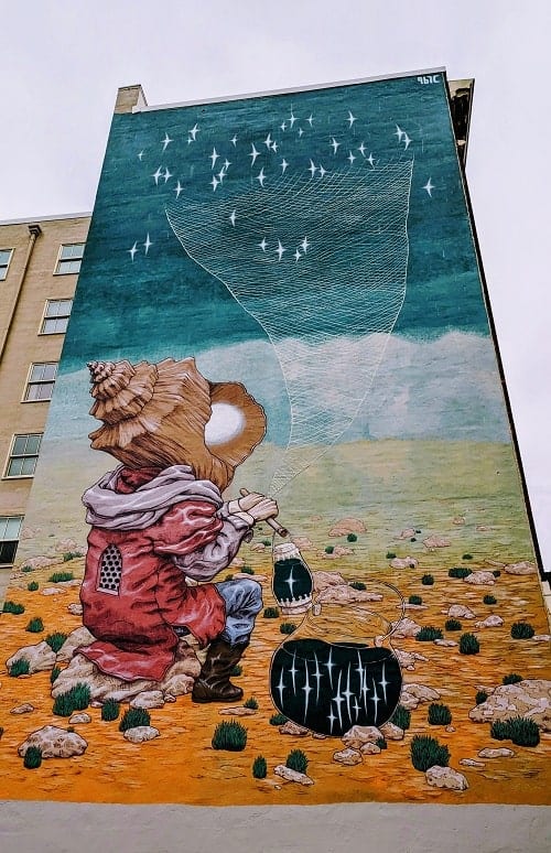 unique mural on the side of a portland building with a small person with a shell head sitting on brown sand creating a tornado