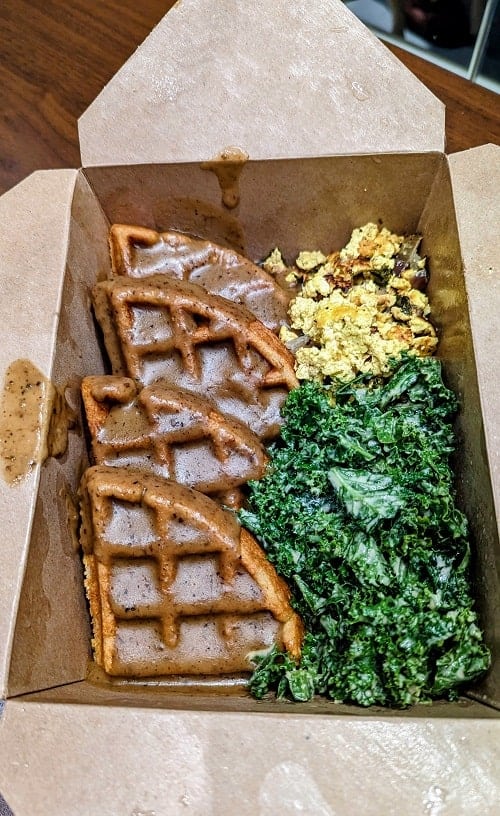 vegan and gluten free golden waffles covered in mushroom rosemary gravy next to tofu scramble and tahini greens in a brown carry out box in portland