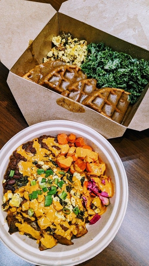vegan and gluten free golden waffles covered in mushroom rosemary gravy next to tofu scramble and tahini greens in a brown carry out box  sitting next to a colorful burrito bowl in portland