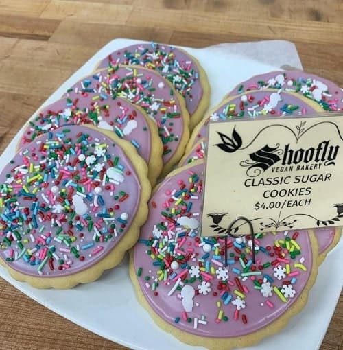 a white plate with six soft baked vegan sugar cookies topped with light purple buttercream and colorful sprinkles from shoo fly bakery in portland