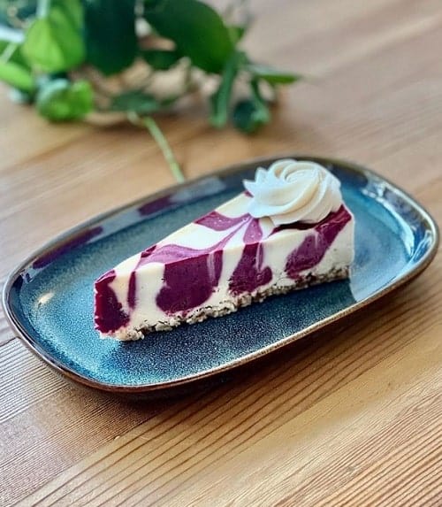 a slice of vegan cheese cake with blueberry swirls on a blue plate sitting on a wood table at moreso in portland