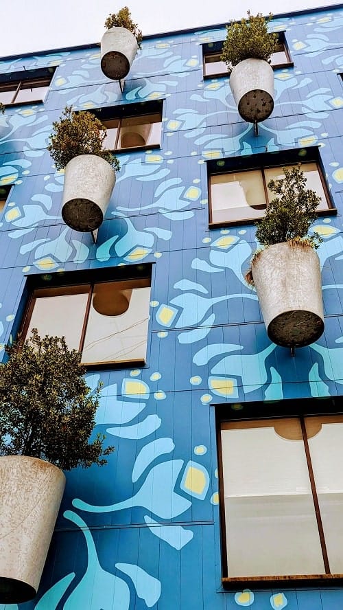 the outside of a blue building with light blue and yellow geometric designs with large silver cylinders filled with mini green trees in portland