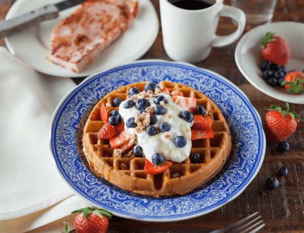 one golden belgian waffle on a blue plate topped with cream and berries next to toast and a white coffee mug