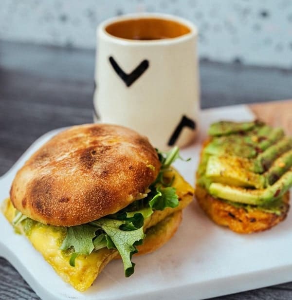 one vegan breakfast sandwich with egg and arugula next to avocado toast and a white coffee mug on a white plate at cafe red in seattle