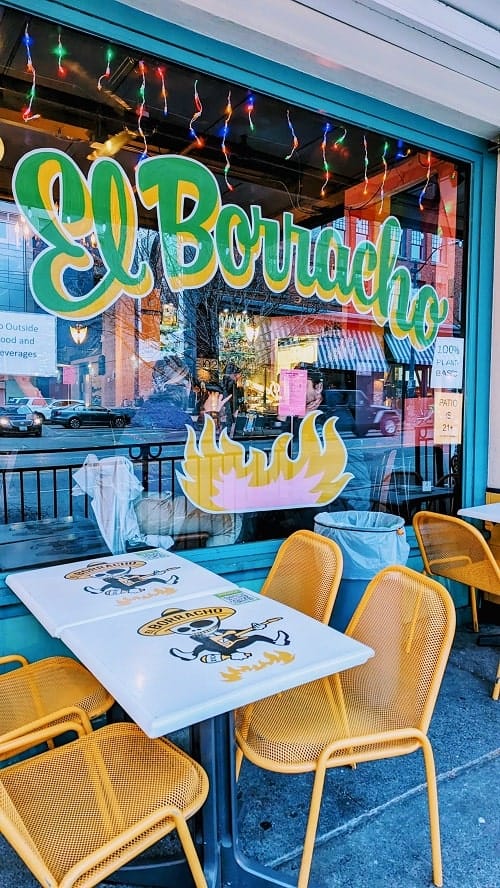the outside of the vegan mexican resturant el borracho with green and yellow lettering and a small white table with four yellow chairs in front in seattle