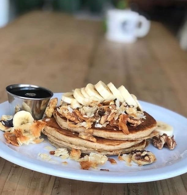 a stack of two golden vegan and gluten free pancakes topped with sliced banana and granola on a white plate at harlow in portland