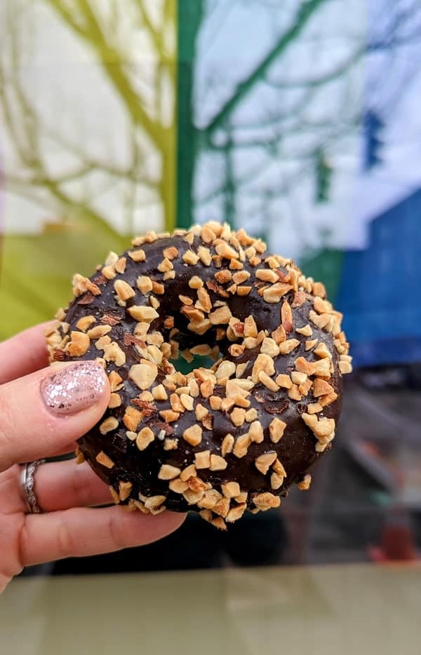 one round vegan chocolate cake donut covered in peanuts held in front of a shop window at mighty o donuts in seattle