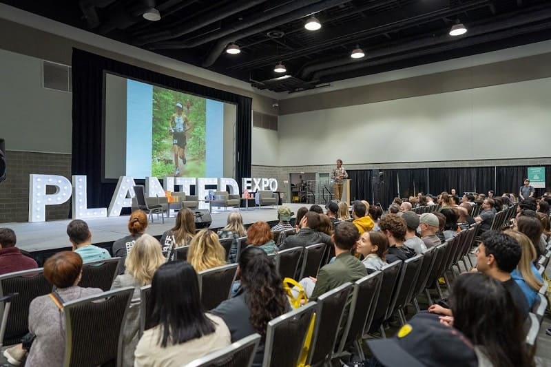 people sitting in chairs in front of a stage with large letters that say planted expo with a man on stage giving a presentation