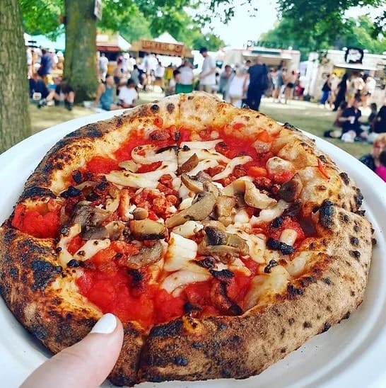 a small vegan pizza held with one hand in front of a lineup of vendors and people in the background