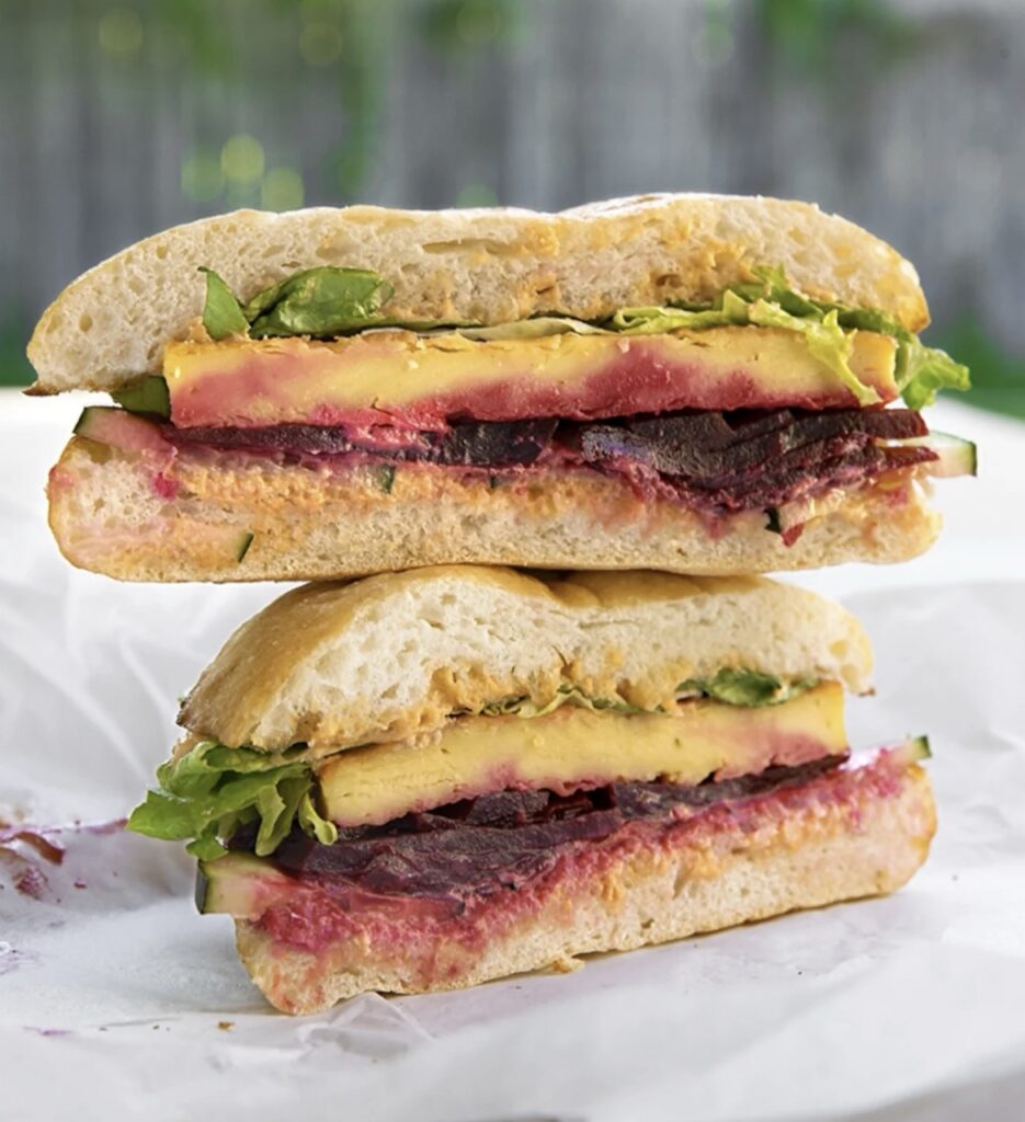 Roast Beet & Panelle Sandwich from Bistro Vonish, featuring smoked beets, golden chickpea cake, tomato aioli, crisp cucumbers, and fresh greens layered between slices of ciabatta bread.