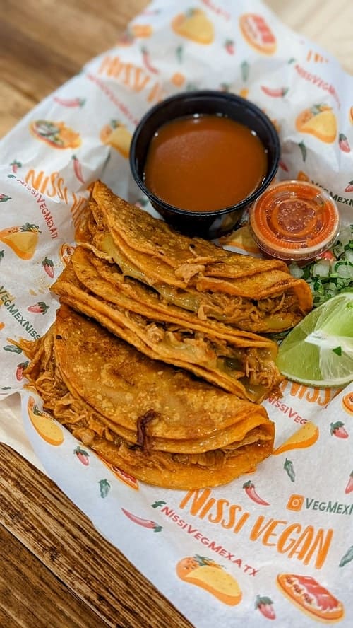 a basket of golden vegan birria tacos next to a cup of dipping sauce at nissi vegmex in austin