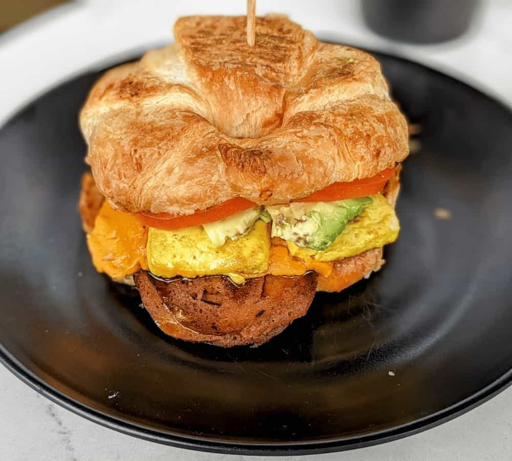 a large vegan croissant breakfast sandwich with egg, tomato, cheese, and bacon on a black plate from rebel cheese in austin