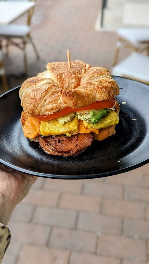 vegan croissant flake and bake breakfast sandwich on a black plate held above a sidewalk at rebel cheese in austin