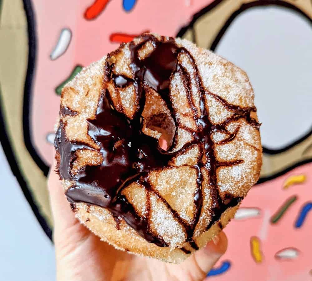 vegan cronut covered in a chocolate drizzle held in front of a pink donut background at machino donuts in toronto