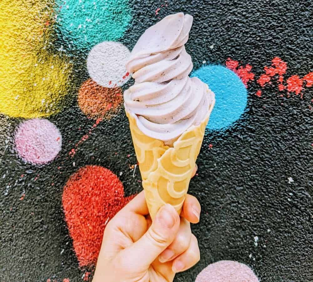 vegan lavender soft serve swirled in a waffle cone and held in front of a black wall with colorful circles at orchard grocer in NYC