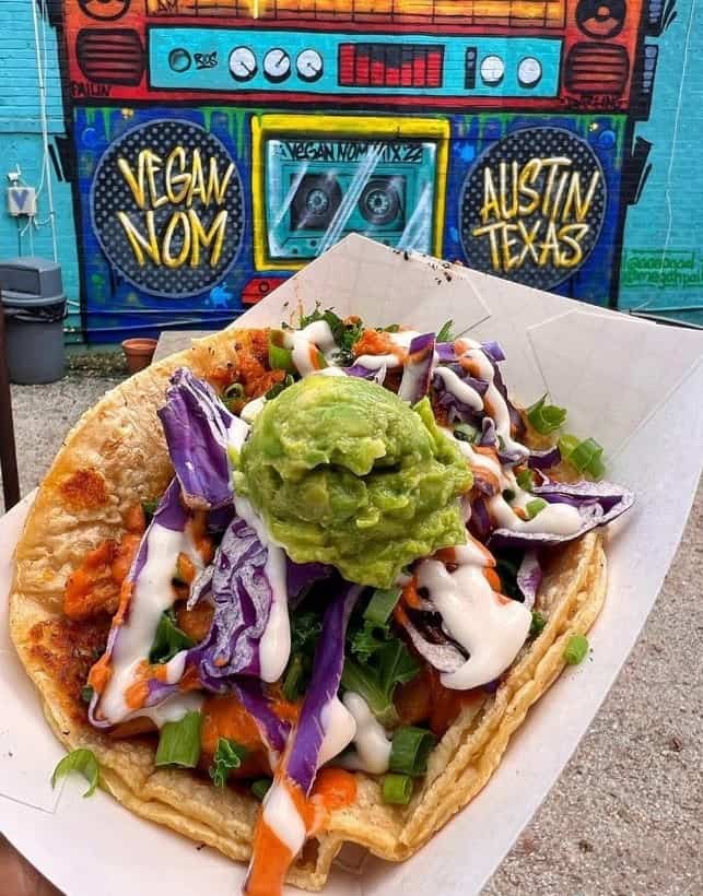 a single vegan taco in a white tray topped with purple cabbage and guacamole held in front of a graffiti mural at the vegan nom in austin