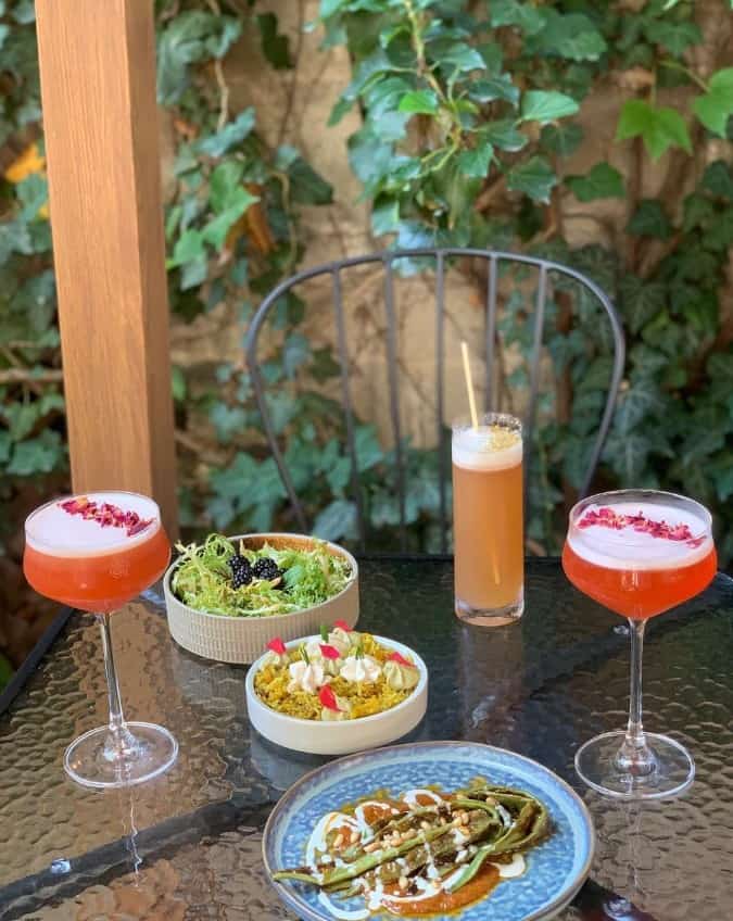 multiple small dishes spread across an outdoor table next to two pink drinks and a peach colored one