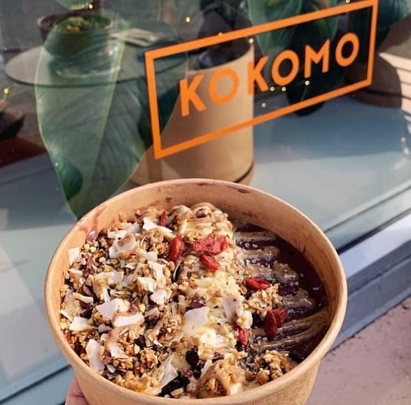 vegan acai bowl topped with fruit, coconut, granola, and a chocolate drizzle held in front of the kokomo sign in vancouver