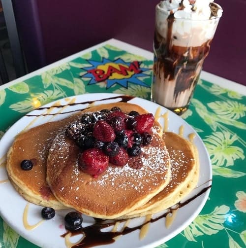 a stack of golden vegan pancakes topped with berries and syrup on a green floral table cloth next to a vegan milkshake at pick me up cafe in chicago