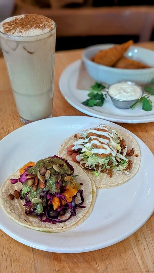 two vegan tacos sitting on a white plate in front of a horchata and chicken wings at the burrow in vancouver