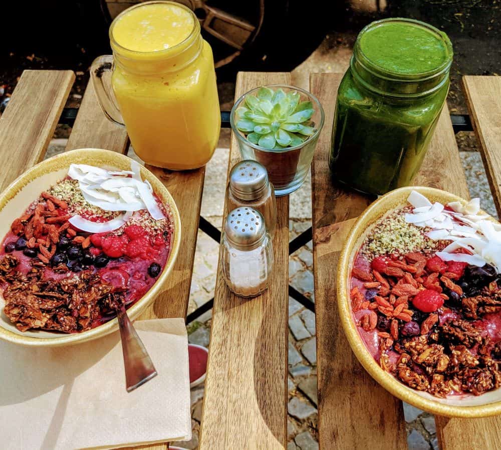 two vegan acai bowls sitting on a wooden table next to a yellow and green smoothie in the summer in berlin