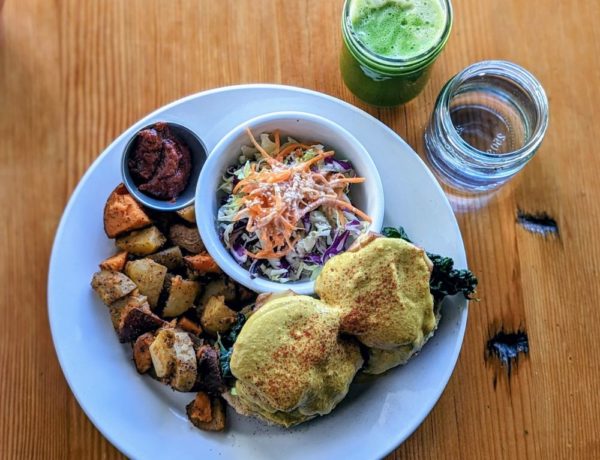 vegan tofu benny breakfast plate with colorful potatoes and slaw next to a green juice and water on a light wood table at eternal abundance in vancouver