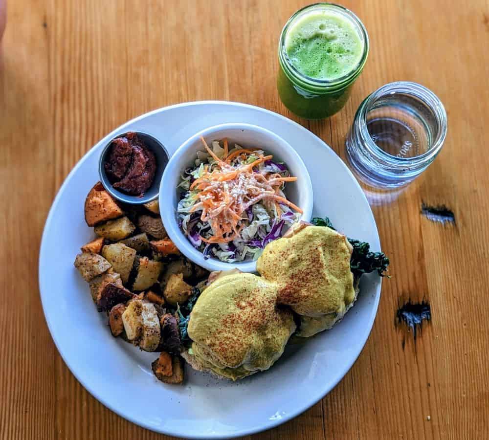 vegan tofu benny breakfast plate with colorful potatoes and slaw next to a green juice and water on a light wood table at eternal abundance in vancouver