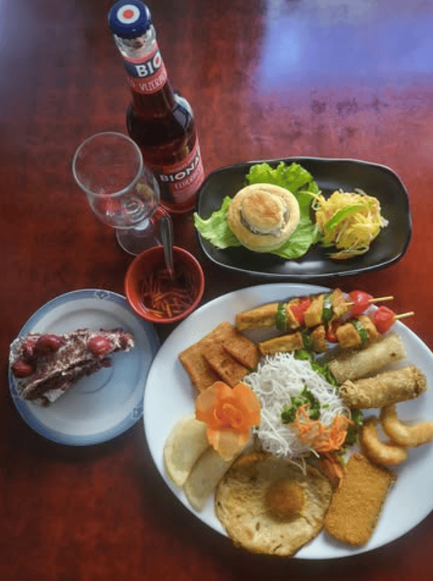 Overhead view of a vegan feast at Bodhi featuring spring rolls, skewers with tofu and vegetables, noodle salad, crispy fried bites, a savory pastry, and a slice of chocolate cake, served alongside a bottle of Bionade on a wooden table.
