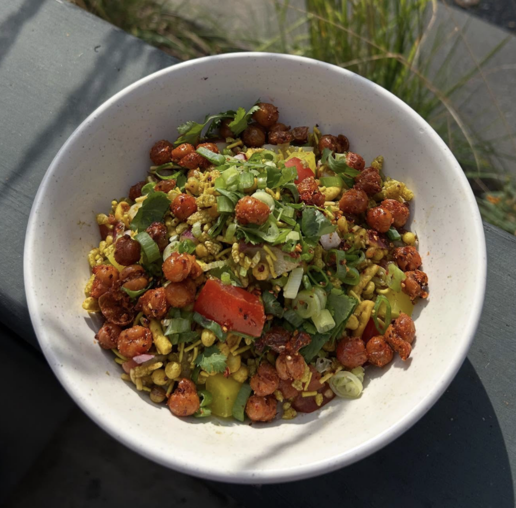 Close-up of a vibrant vegan Bombay bhel bowl topped with crispy chickpeas, chopped herbs, tomatoes, and crunchy puffed grains, tossed with chutneys and served in a white bowl outdoors.