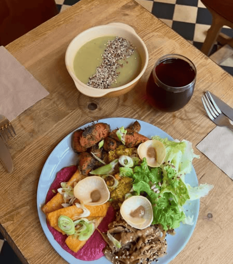 Overhead view of a colorful vegan meal at Sol Semilla featuring roasted vegetables, fresh greens, mushrooms, beet purée, and crispy chips served on a blue plate, alongside a creamy soup topped with seeds and a dark herbal drink on a wooden table.