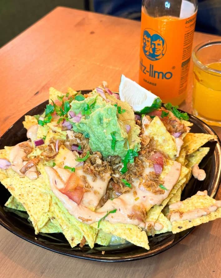 a giant plate of vegan nachos topped with cheese, guac, and more. next to a glass bottle of pop at plant base in lisbon