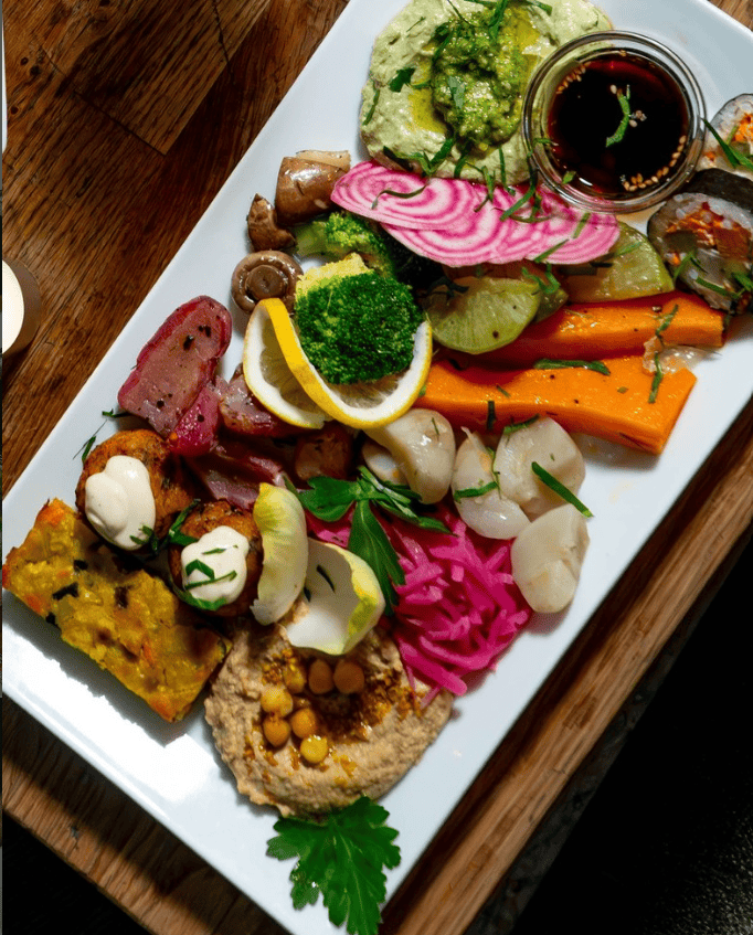 Overhead view of a colorful vegan platter at Soya featuring hummus topped with chickpeas, pickled vegetables, roasted carrots, broccoli, mushrooms, stuffed sushi rolls, falafel with creamy sauce, and assorted Mediterranean-inspired small bites served on a large white plate.