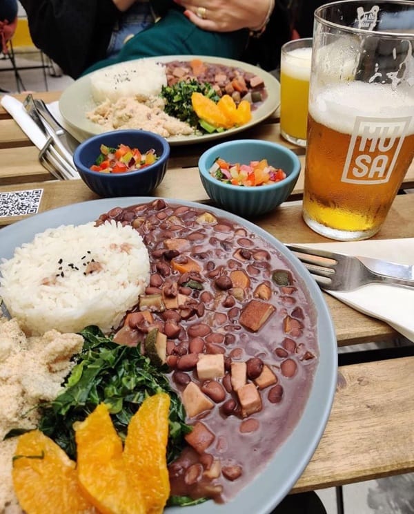 an up close shot of a large round bowl filled with dark beans, rice, and plantains on an outdoor table next to a beer thats been half drunk and another bowl in the background